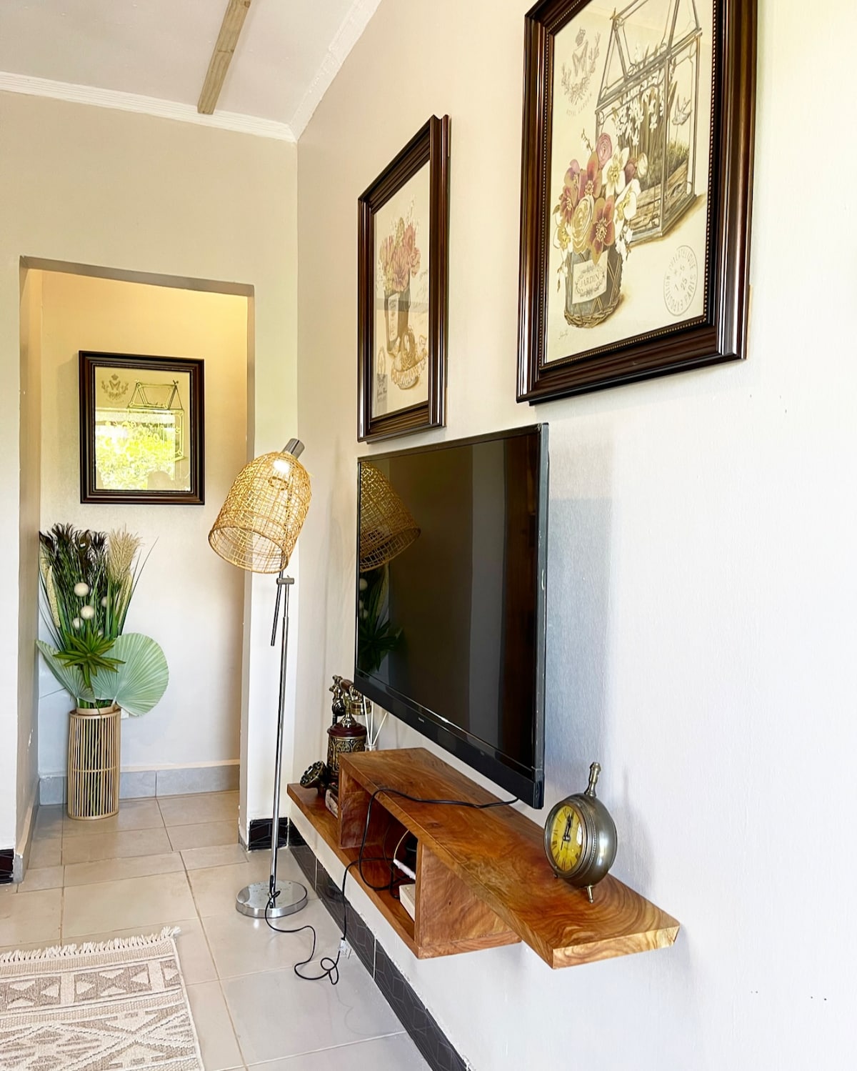 A wall-mounted television is displayed above a wooden shelf. Decorative frames featuring floral artwork adorn the walls. A stylish floor lamp stands nearby, accompanied by a decorative vase with plants, adding natural elements to the bright space.