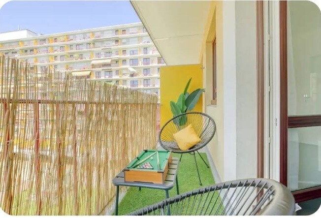 A balcony is depicted, featuring two stylish chairs and a small table with a game set. A green plant adds a touch of nature, and a bamboo screen provides privacy. The backdrop shows a building facade with colorful accents, enhancing the outdoor setting.