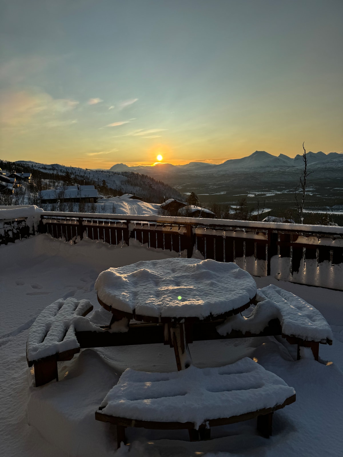 A round wooden table is surrounded by benches, all covered in fresh snow. The space is illuminated by a soft sunset, with mountains in the distance appearing under a colorful sky. The tranquil atmosphere is enhanced by the winter landscape surrounding the area.