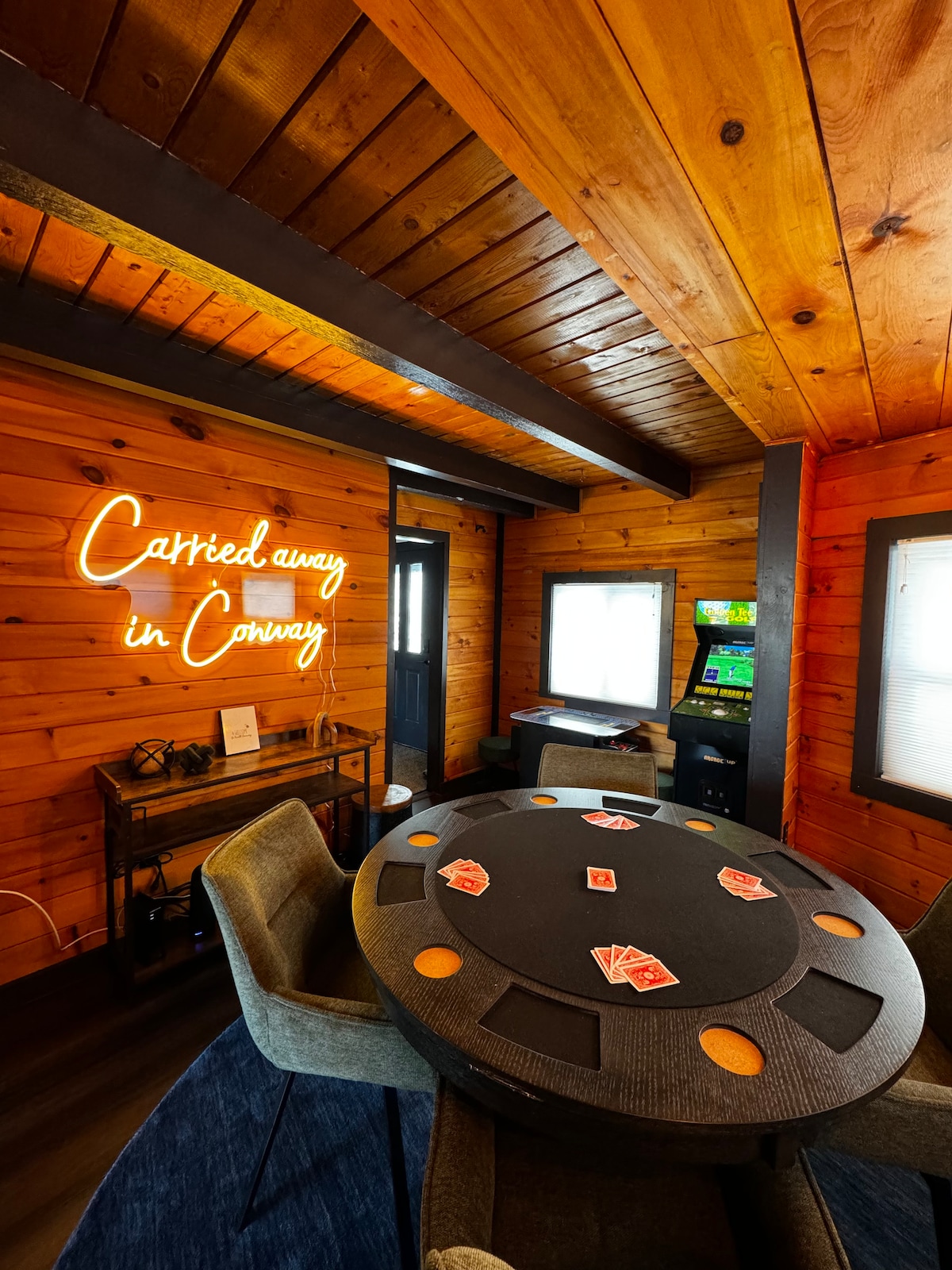 A cozy entertainment area features warm wooden walls and a circular poker table, surrounded by upholstered chairs. A neon sign showcasing 'Carried away in Conway' is prominently displayed. An arcade game machine is visible in the background, contributing to the recreational atmosphere.