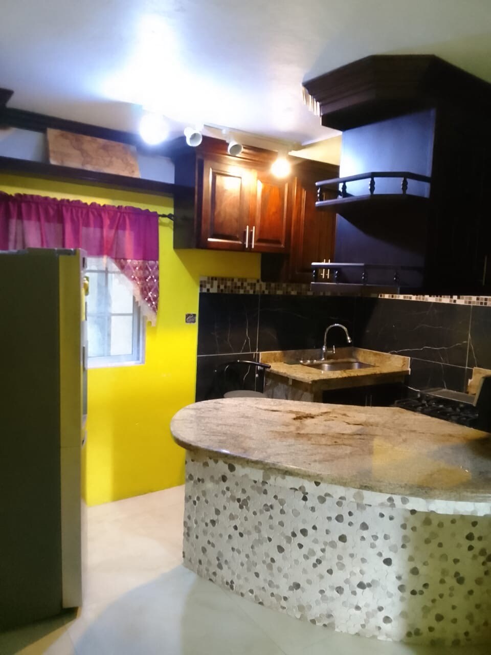 A modern kitchen is highlighted with dark wooden cabinets and a large stone countertop. Bright yellow walls contrast with dark accents, and overhead lighting illuminates the space. A spacious sink is positioned near the countertop, while a refrigerator stands against the wall.