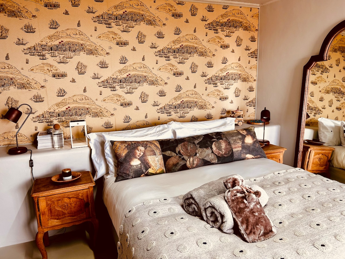 A spacious bed is presented with patterned pillows and a soft throw, complemented by side tables featuring lamps and decor. The neutral-toned walls are adorned with a nautical-themed wallpaper, adding character to the room. A mirror reflects the inviting space, enhancing natural light.