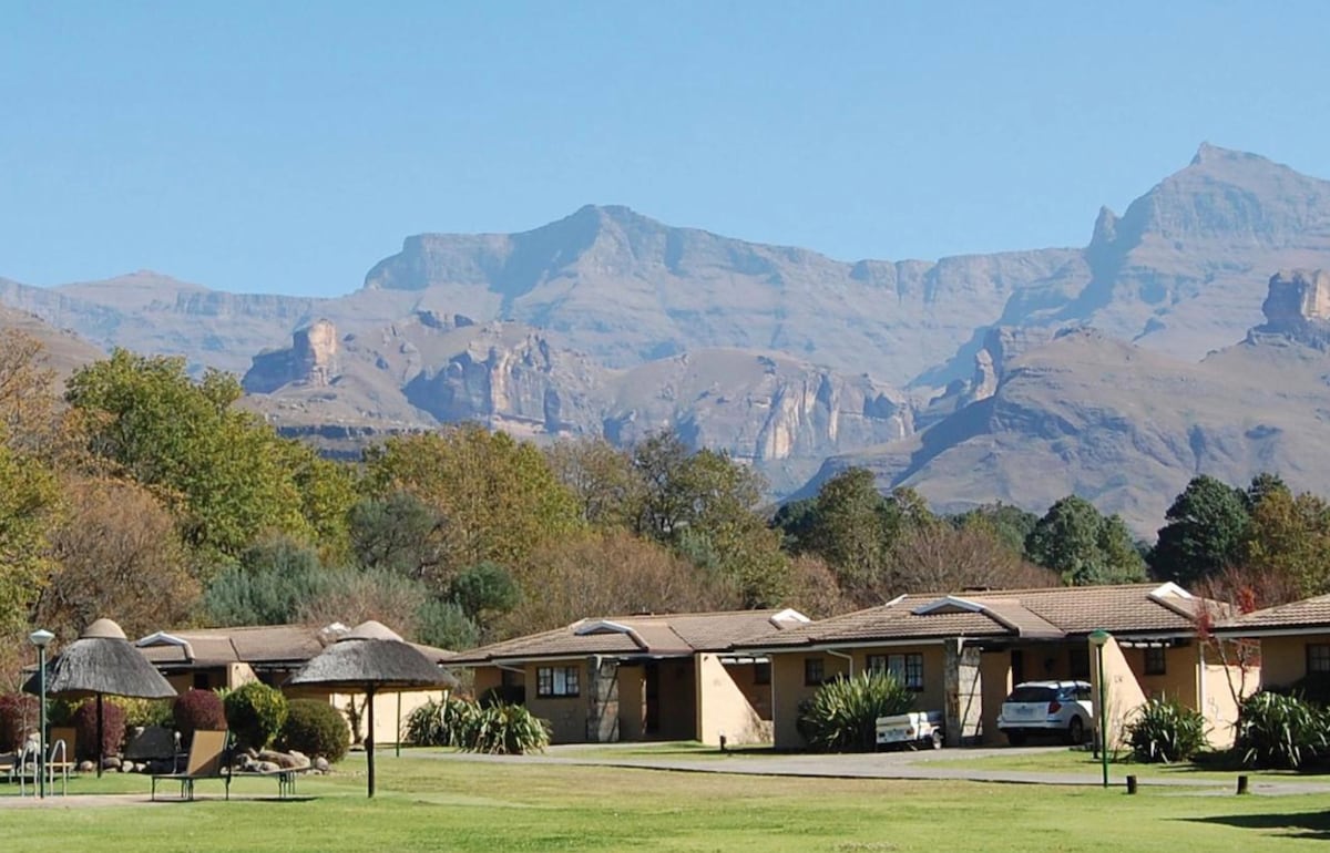 The image captures the scenic landscape of the Drakensberg Mountains, serving as a backdrop to the resort's chalets. Lush greenery surrounds the area, while thatched roofs of the chalets blend harmoniously with the natural environment. Clear blue skies contribute to the serene atmosphere.