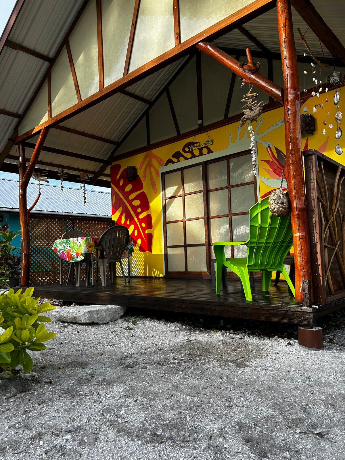 A terrace deck is featured in this image, showcasing a colorful table with floral patterns and two chairs, one in vibrant green. The wall behind displays bold tropical-inspired artwork, while a stone pathway leads to the entrance of the bungalow.