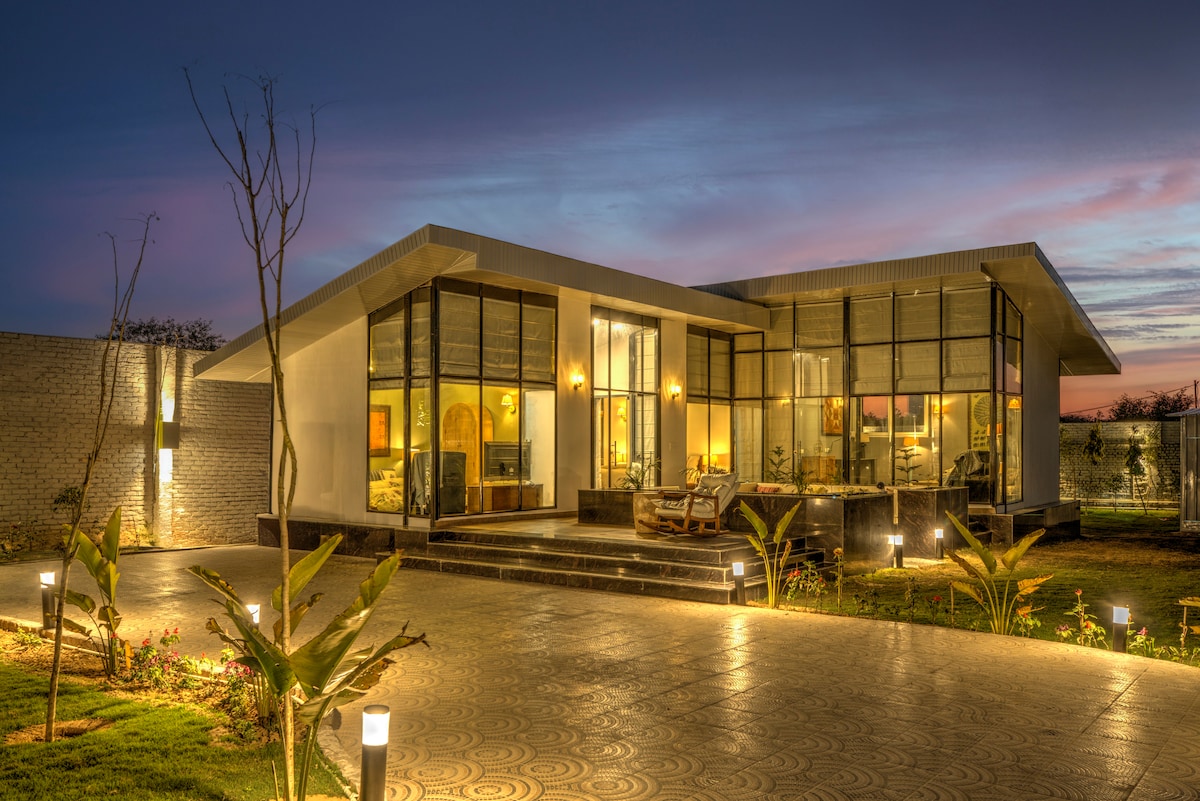 The exterior of a modern house is showcased at dusk, illuminated by soft lighting. The structure features large glass windows and a sleek design, surrounded by landscaped gardens. Pathway lights add a warm glow, enhancing the inviting atmosphere of the property.