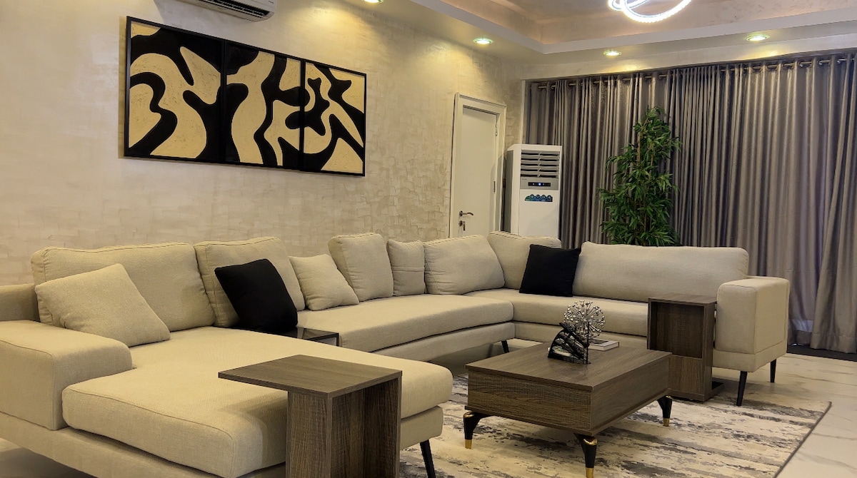 A spacious living area features a light-colored sectional sofa with accent pillows and two wooden side tables. A stylish coffee table rests at the center, complemented by a decorative item. Floor-to-ceiling curtains frame large windows, while a plant adds a touch of greenery.