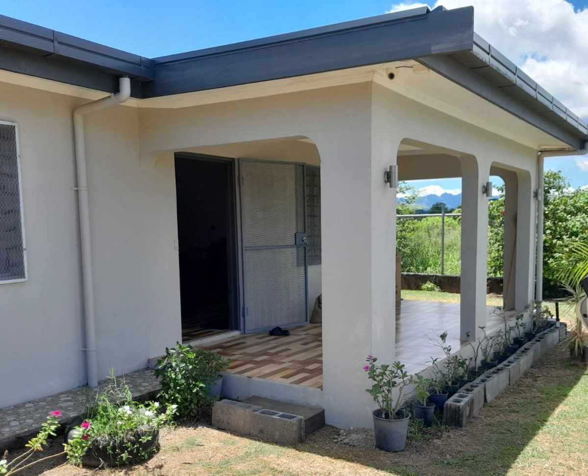 The exterior of the apartment showcases a covered porch with tiled flooring. Potted plants line the edge, contributing to the greenery surrounding the property. The entrance is framed by a simple structure, with a screened door providing access to the interior.