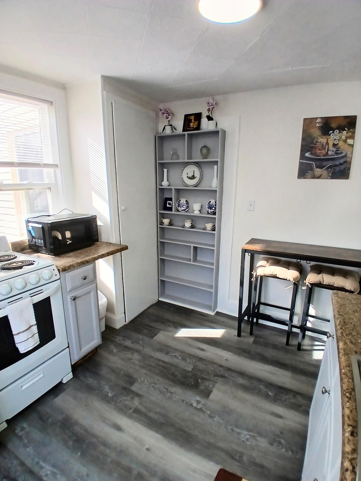 A modern kitchen space features a stylish stove and microwave, complemented by a bar-height table with stools. Natural light illuminates the area through a window, while a shelf displays decorative items, enhancing the functional aspect of the space.