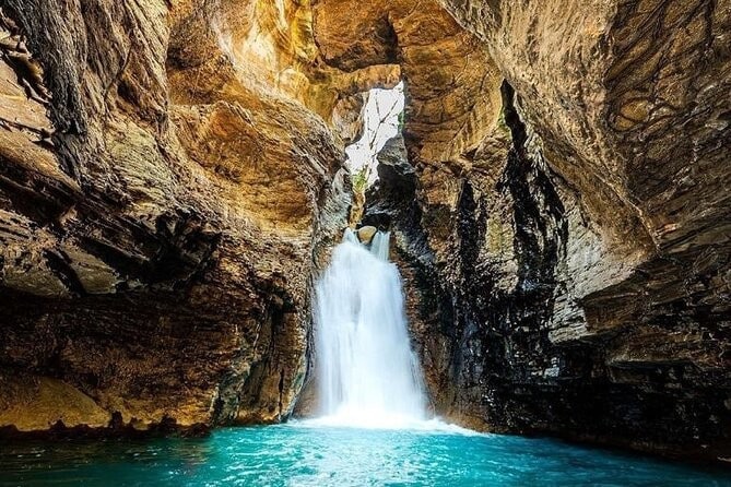 A waterfall cascades over rocky formations into a serene turquoise pool, framed by rugged stone walls. The natural light filters through the openings above, creating a soft glow on the water's surface, enhancing the tranquil ambiance of this secluded spot.