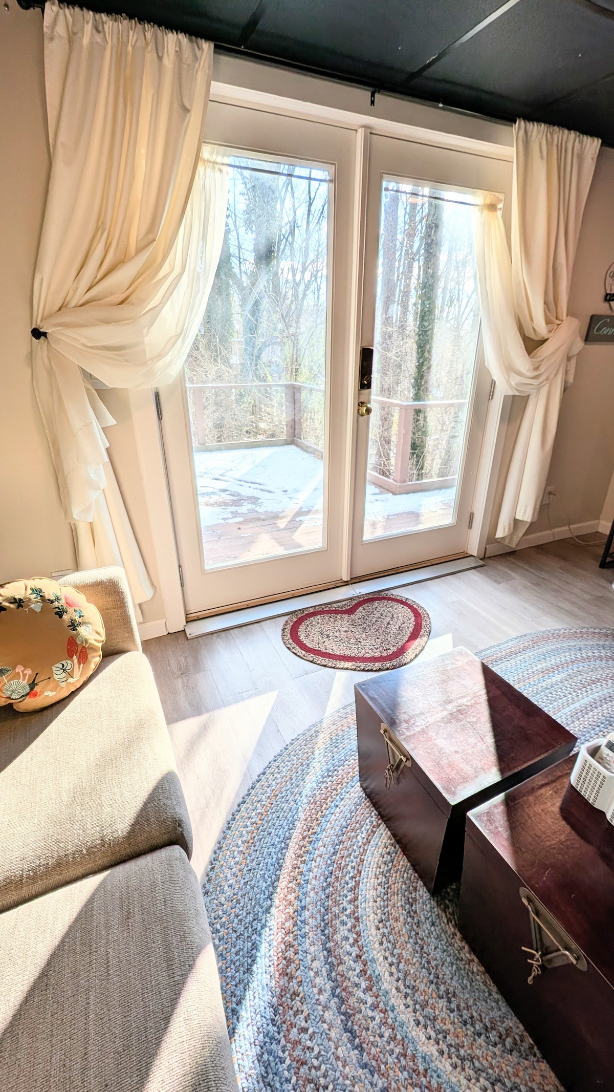 Natural light fills the space through glass doors, leading to a private deck. Sheer curtains frame the entrance, while a small couch and a round area rug enhance the cozy atmosphere. A heart-shaped mat is positioned by the doors, adding a touch of warmth.