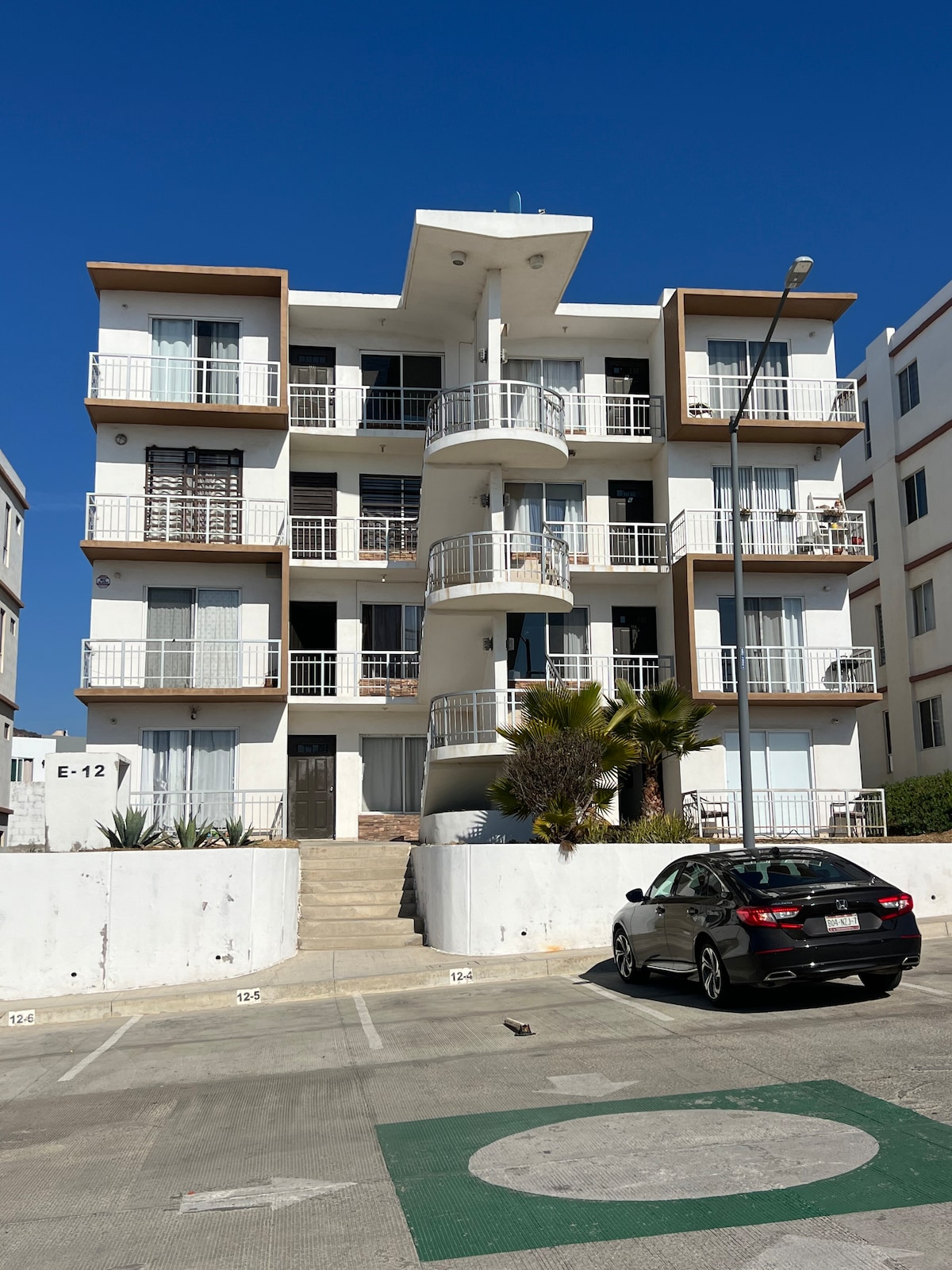 A modern three-story building features multiple balconies with railings and large windows, allowing ample natural light. The entrance is accessed via a staircase surrounded by landscaped greenery. A car is parked in the foreground, indicating convenient parking options.
