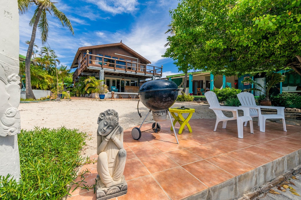 A charming outdoor area is presented, featuring a sandy space surrounded by tropical greenery. A charcoal grill sits on tiled flooring beside a white adirondack chair, while a decorative statue adds character to the setting. The background reveals a two-story house and additional seating areas.