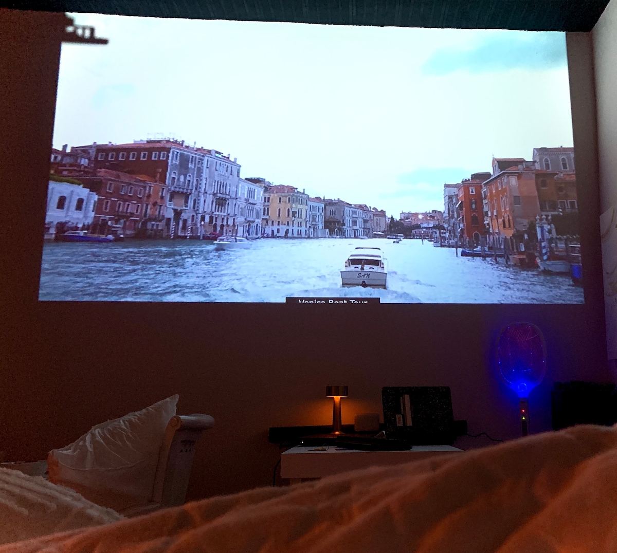 A large projection of a scenic waterway with boats can be seen on the wall, creating a cinematic atmosphere. In the foreground, a cozy bed is lightly visible, with soft pillows and a textured blanket. The room is dimly lit, adding to the immersive experience.