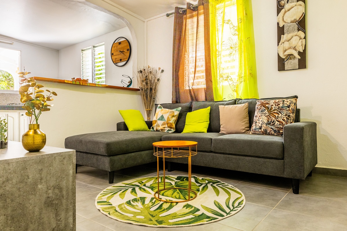 A modern living area is featured, showcasing a gray sectional sofa adorned with pillows in various colors and patterns. A circular rug with a leafy design is placed under a small, round gold table. Natural light filters through bright curtains, highlighting the space's inviting ambiance.
