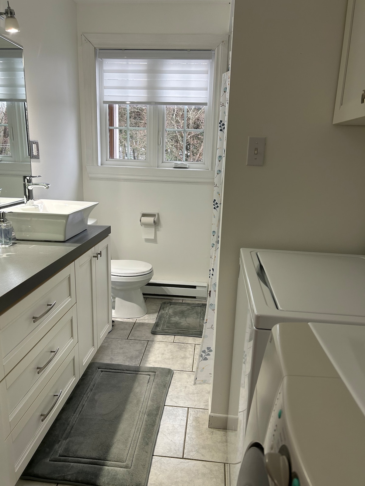 A well-organized bathroom is highlighted by modern fixtures and a clean design. A white sink is positioned on a countertop, with a spacious mirror above. Natural light enters through a window, which is adorned with blinds, enhancing the overall brightness of the space.