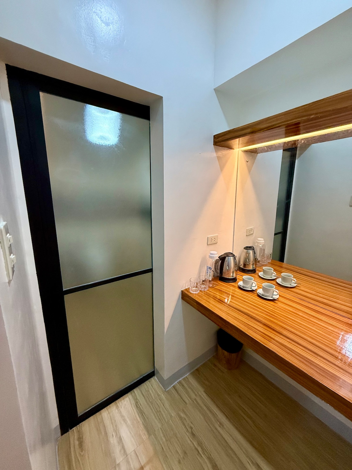 A small kitchenette area is shown with a wooden countertop displaying tea and coffee-making supplies, including a kettle and cups. A frosted glass door leads to another space, while a mirror with subtle lighting enhances the area. Light-colored wood flooring is visible.
