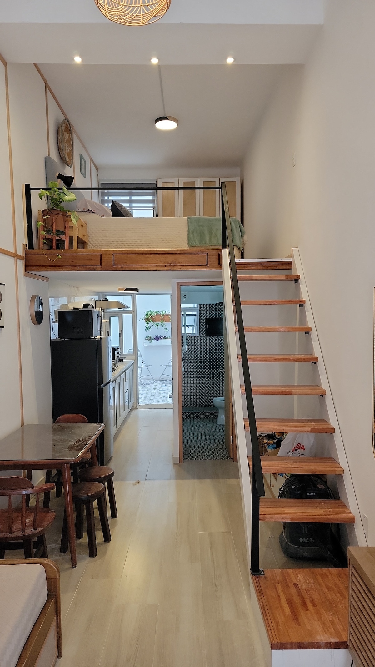 An open-plan living area features a staircase leading to a lofted sleeping space. A dining table with chairs is present on the left, while an equipped kitchen is visible in the background. Natural light illuminates the space through multiple sources.