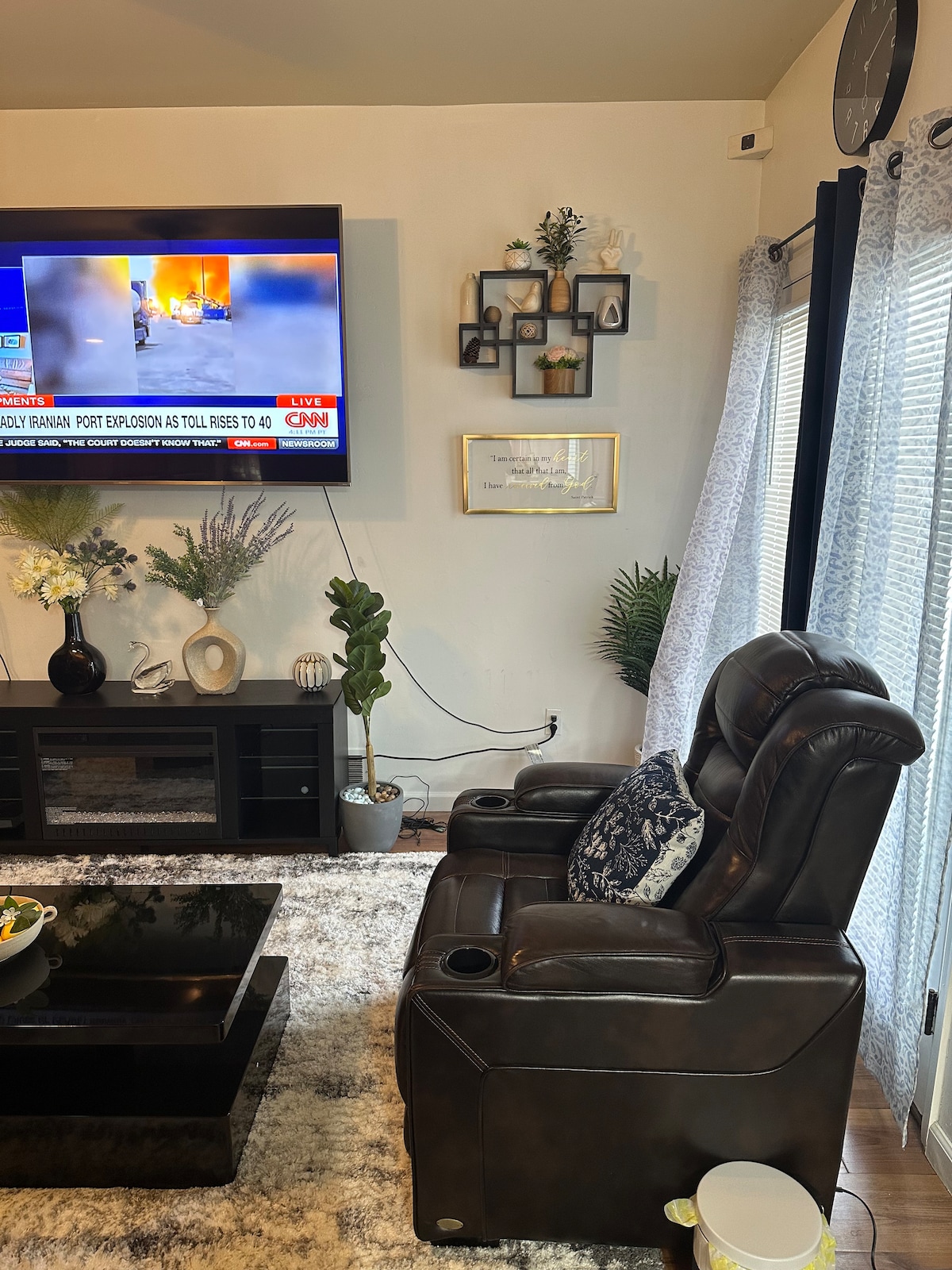 A comfortable reclining chair sits in front of a large television, showcasing news coverage. A decorative wall with framed photos and plants adds visual interest. Sheer curtains allow light to filter through, enhancing the cozy ambiance of the living area.