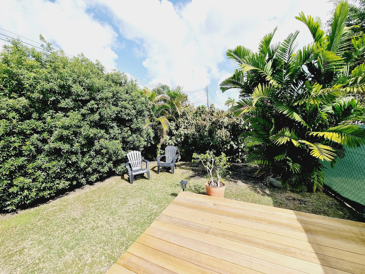 A private outdoor garden features a wooden deck with two gray Adirondack chairs positioned among lush greenery. Various tropical plants are visible throughout the space, creating a tranquil environment. The sky is bright with scattered clouds, enhancing the openness of the area.
