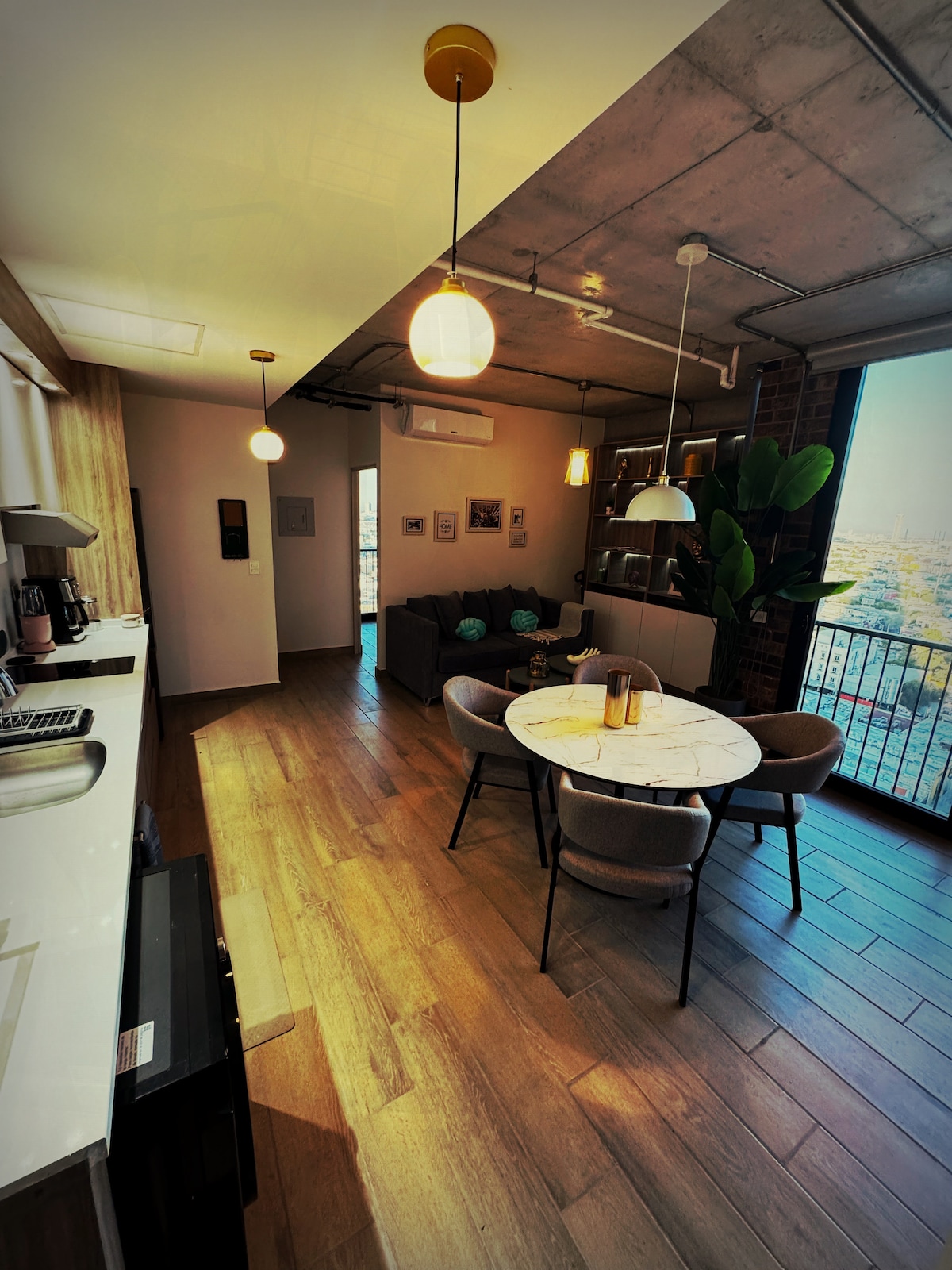 A contemporary open-concept space is featured, with a modern kitchen featuring a coffee maker and a dark countertop. A round dining table with four upholstered chairs is positioned beside a large window, allowing natural light to illuminate the living area, which includes a comfortable sofa and decorative plants.