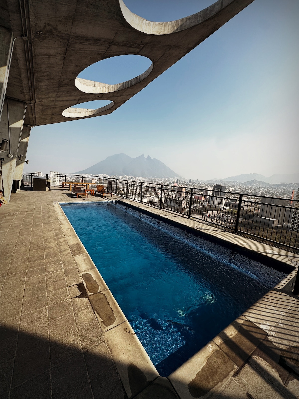 An outdoor pool is situated on a terrace, offering panoramic views of the surrounding city and mountains. The pool's clear blue water reflects the sky, while a few lounge chairs are positioned nearby for relaxation. The concrete structure overhead features circular cutouts, providing an architectural element.