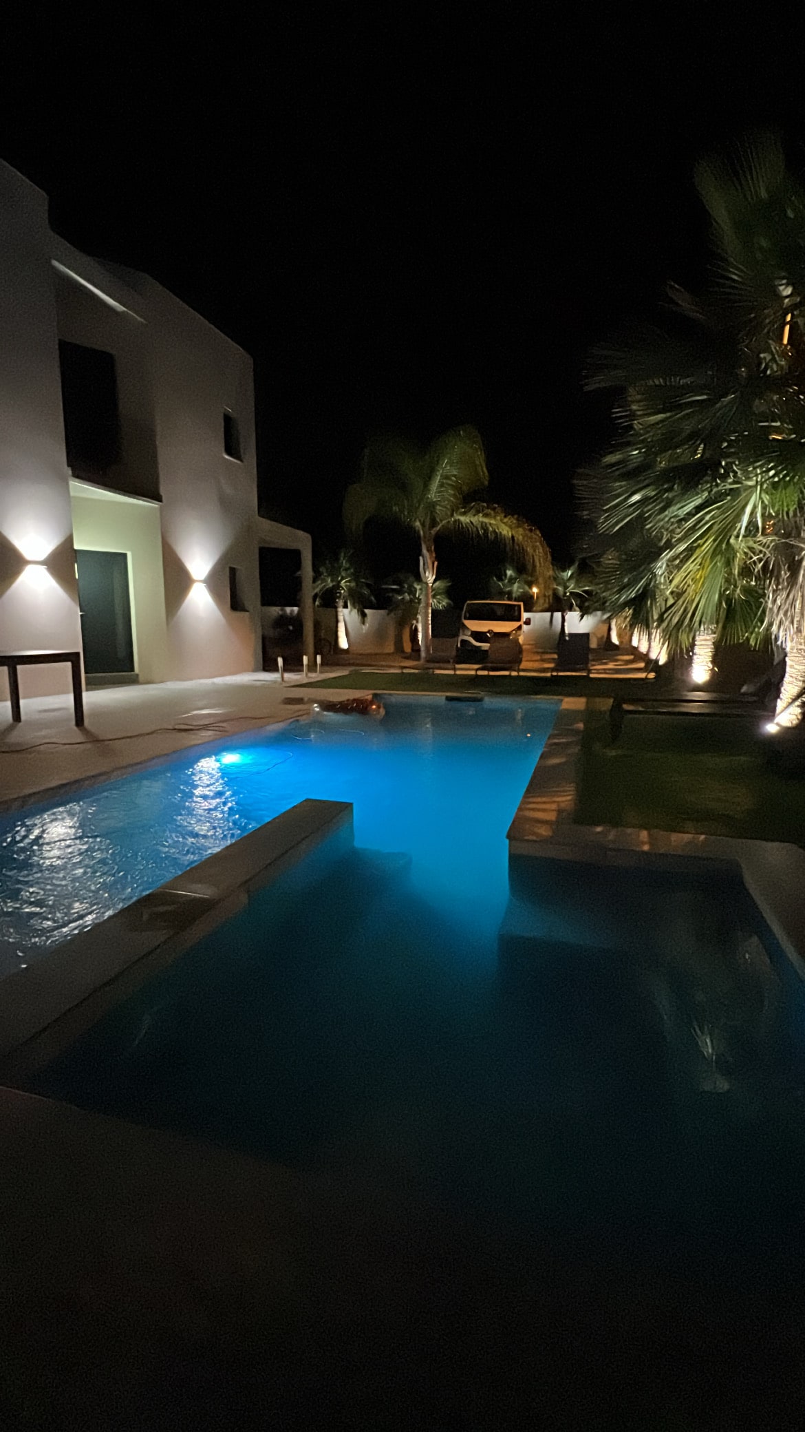 A private pool is illuminated at night, showcasing soft blue tones amidst a serene setting. The surrounding area features palm trees and well-placed lighting, enhancing the tranquil atmosphere. The home's architecture is visible in the background, providing a modern contrast to the natural surroundings.