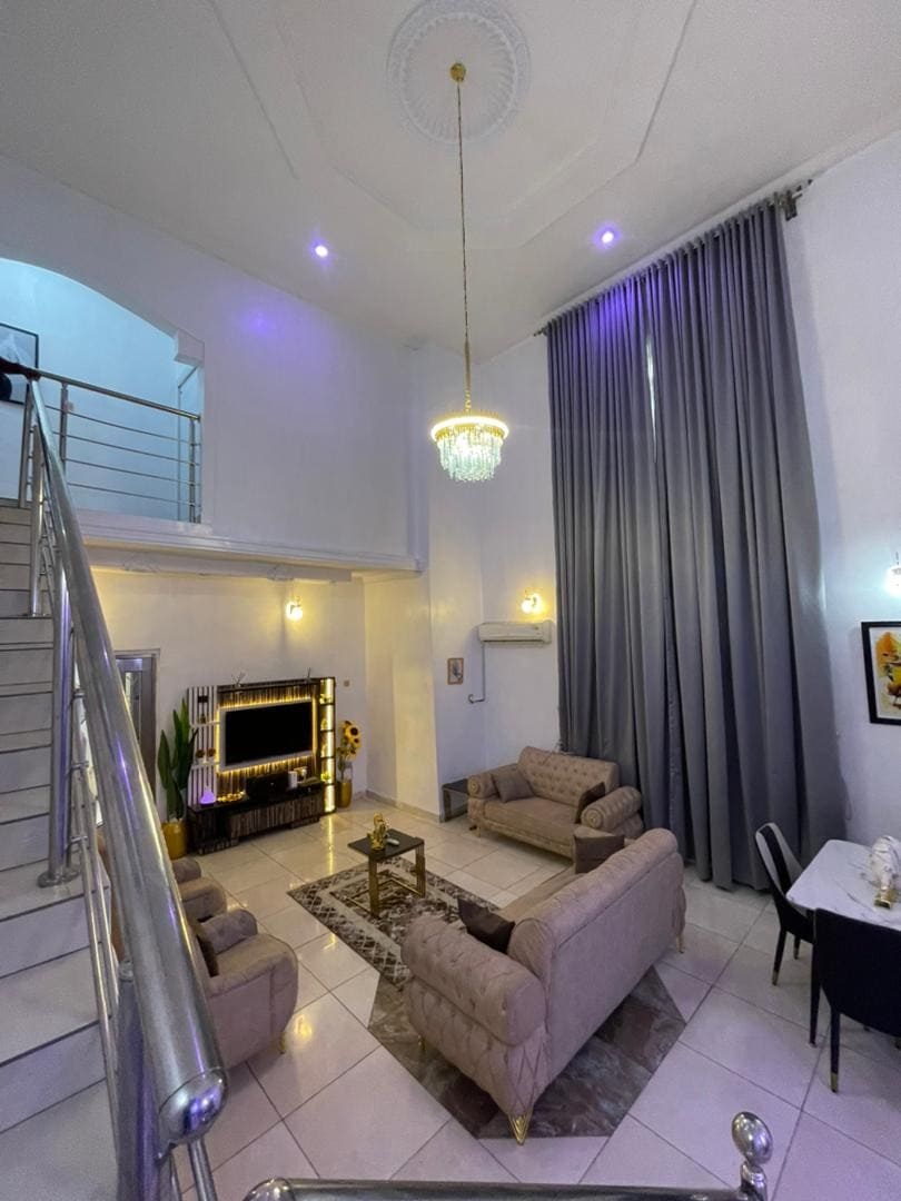 A spacious living area features two plush sofas facing a modern fireplace, with a glass coffee table placed in between. Tall curtains frame the large windows, and a decorative chandelier adds elegance to the high ceiling. A dining area is visible in the background.