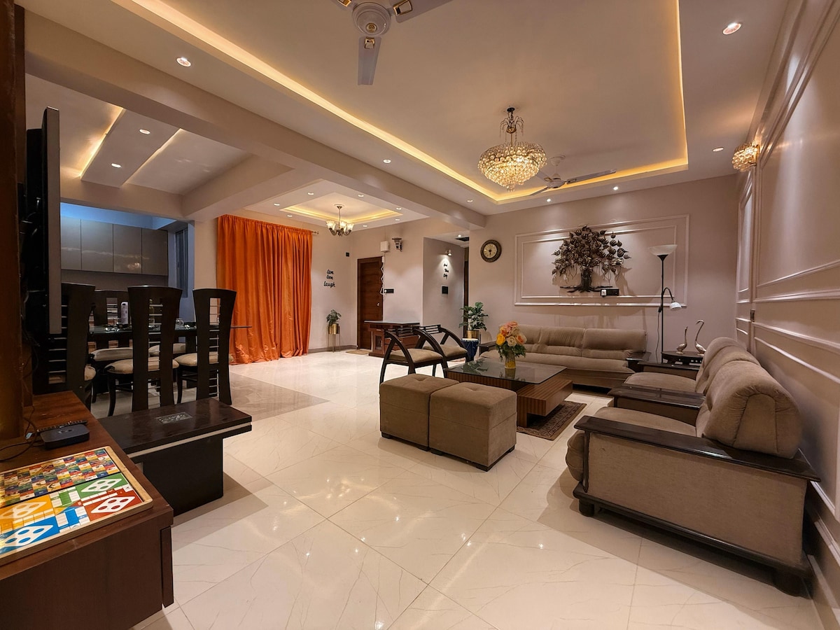 An elegant living area is showcased with soft seating arranged around a coffee table, complemented by a dining set in the background. A warm orange curtain drapes across one wall, while soft lighting highlights the modern decor and gleaming floor tiles.