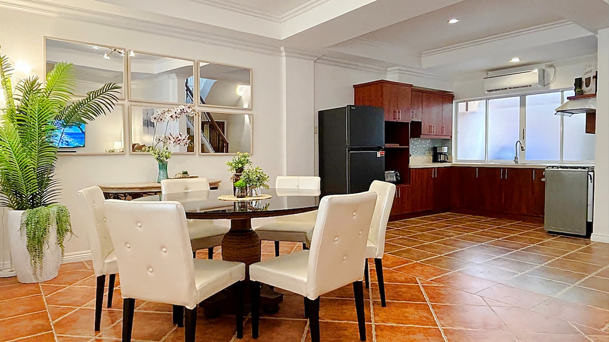 The open-concept living and dining area showcases a round dining table surrounded by six white chairs. A modern kitchen is visible in the background, featuring dark wood cabinetry and stainless-steel appliances. Light floods the space through large windows, enhancing the inviting atmosphere.