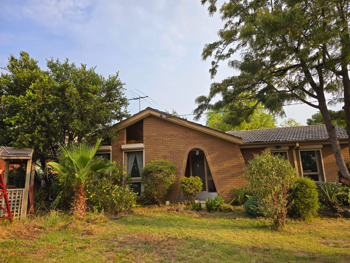The exterior of a single-story brick home is showcased, framed by lush greenery and gardens. A unique arched entryway leads into the home, while palm and other plants add a natural touch to the inviting landscape.