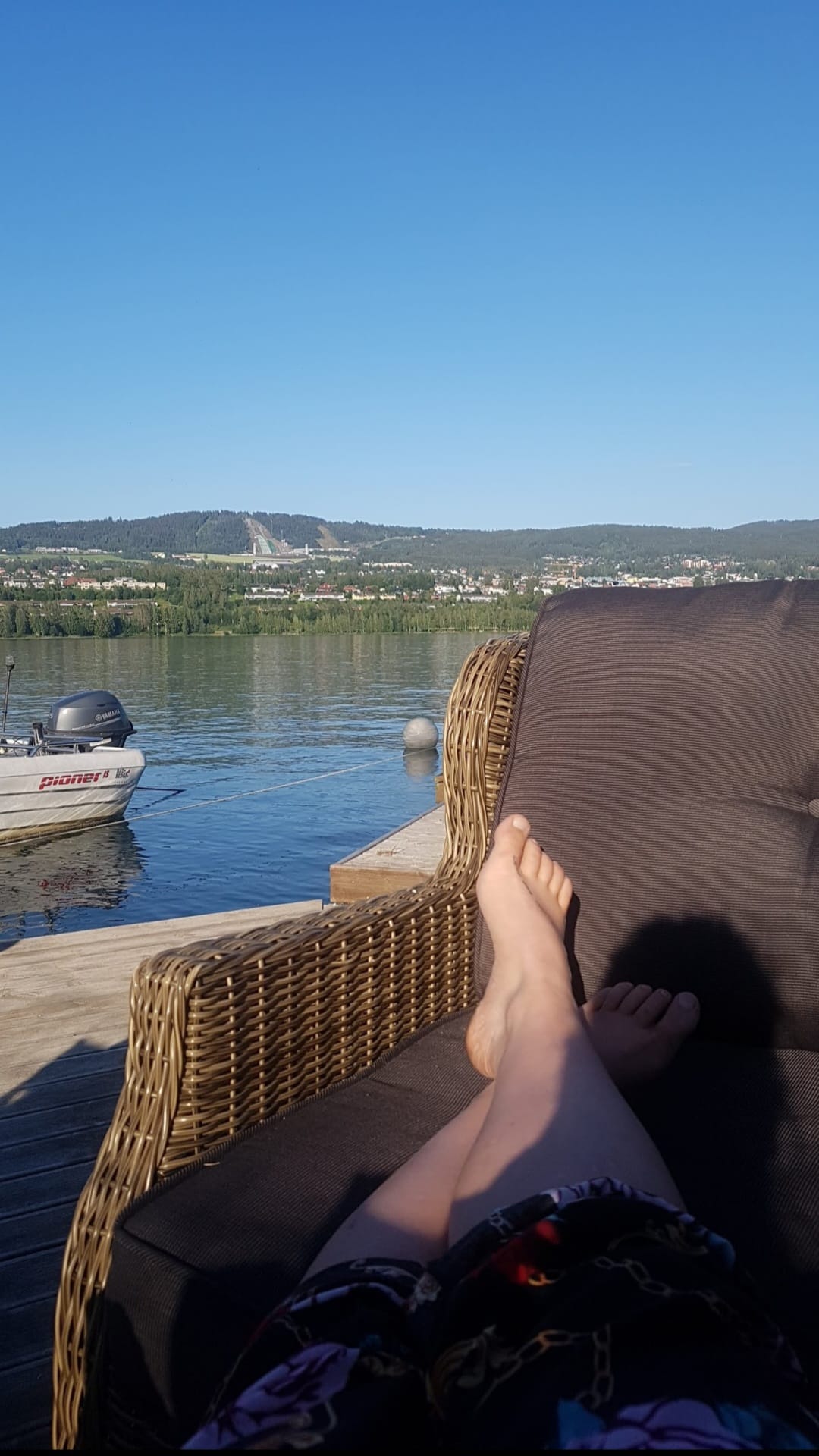 A comfortable wicker chair is positioned on a wooden deck overlooking a calm body of water. Bare feet rest on the chair, with a boat floating nearby. The hills and cityscape of Lillehammer are visible in the background under a clear blue sky.