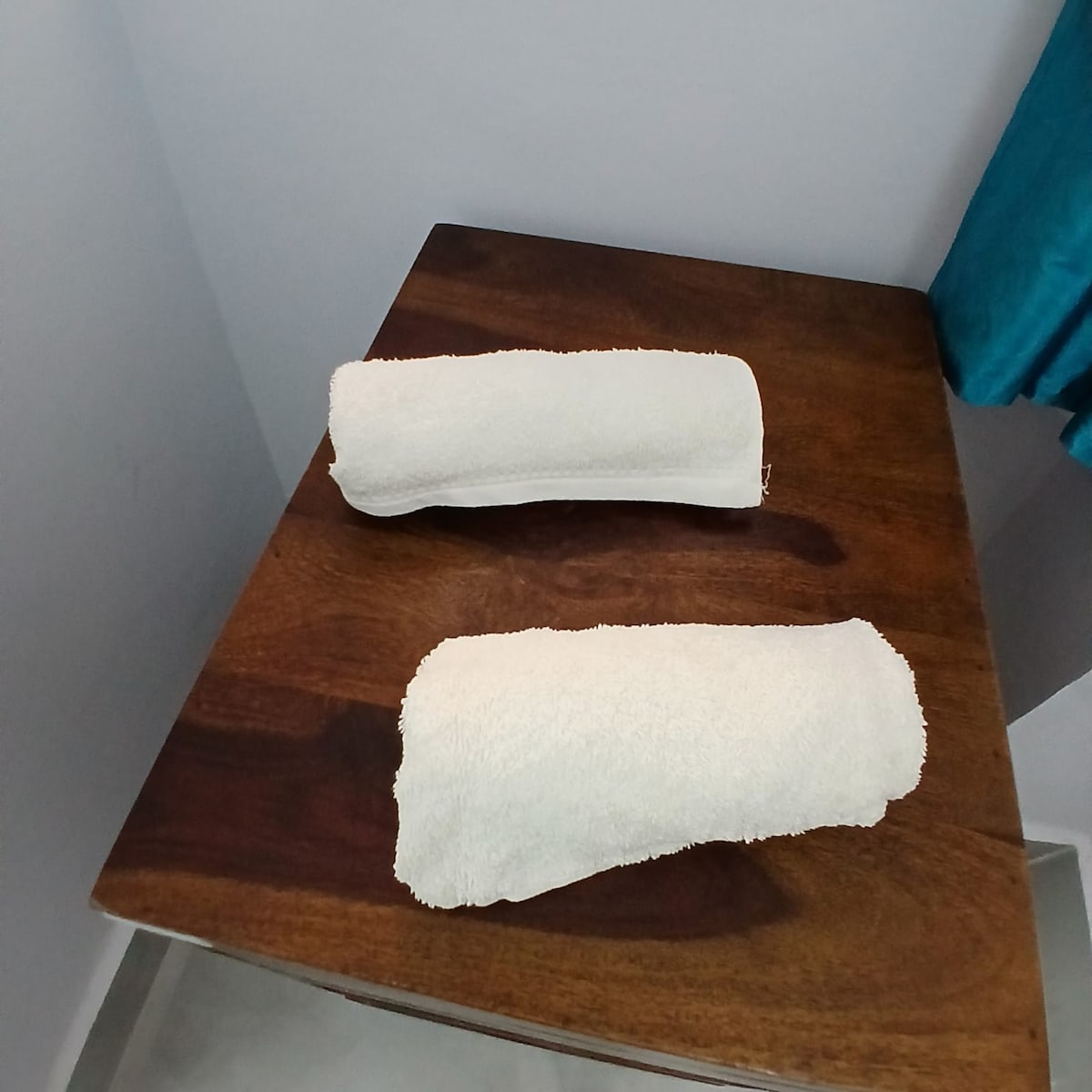 Two neatly rolled white towels are placed on a wooden surface. The soft, textured fabric contrasts with the dark grain of the table. A hint of turquoise fabric is visible in the background, adding a pop of color to the serene setting.