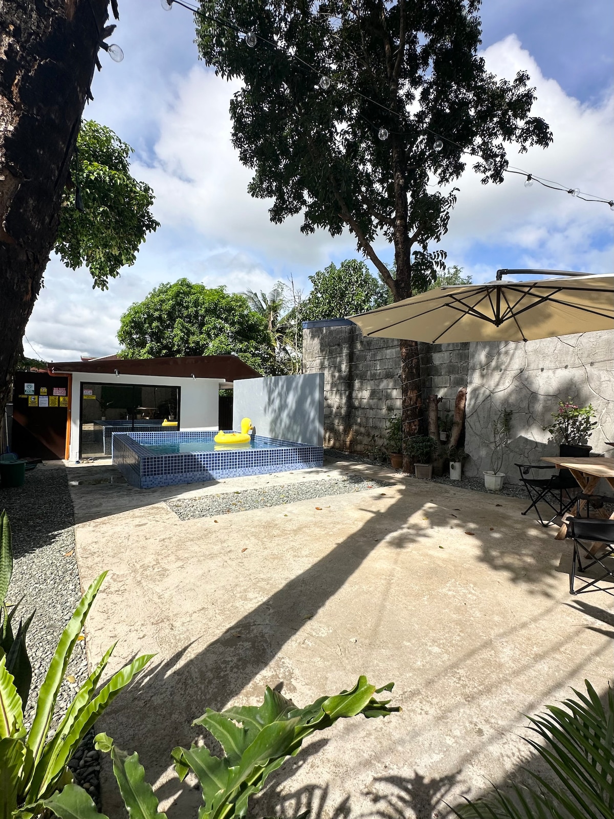 A private outdoor space features a rectangular swimming pool surrounded by a textured stone patio. Tall trees provide shade, while a sun umbrella offers additional coverage. A small dining setup with chairs and a table is situated nearby, complemented by lush greenery.