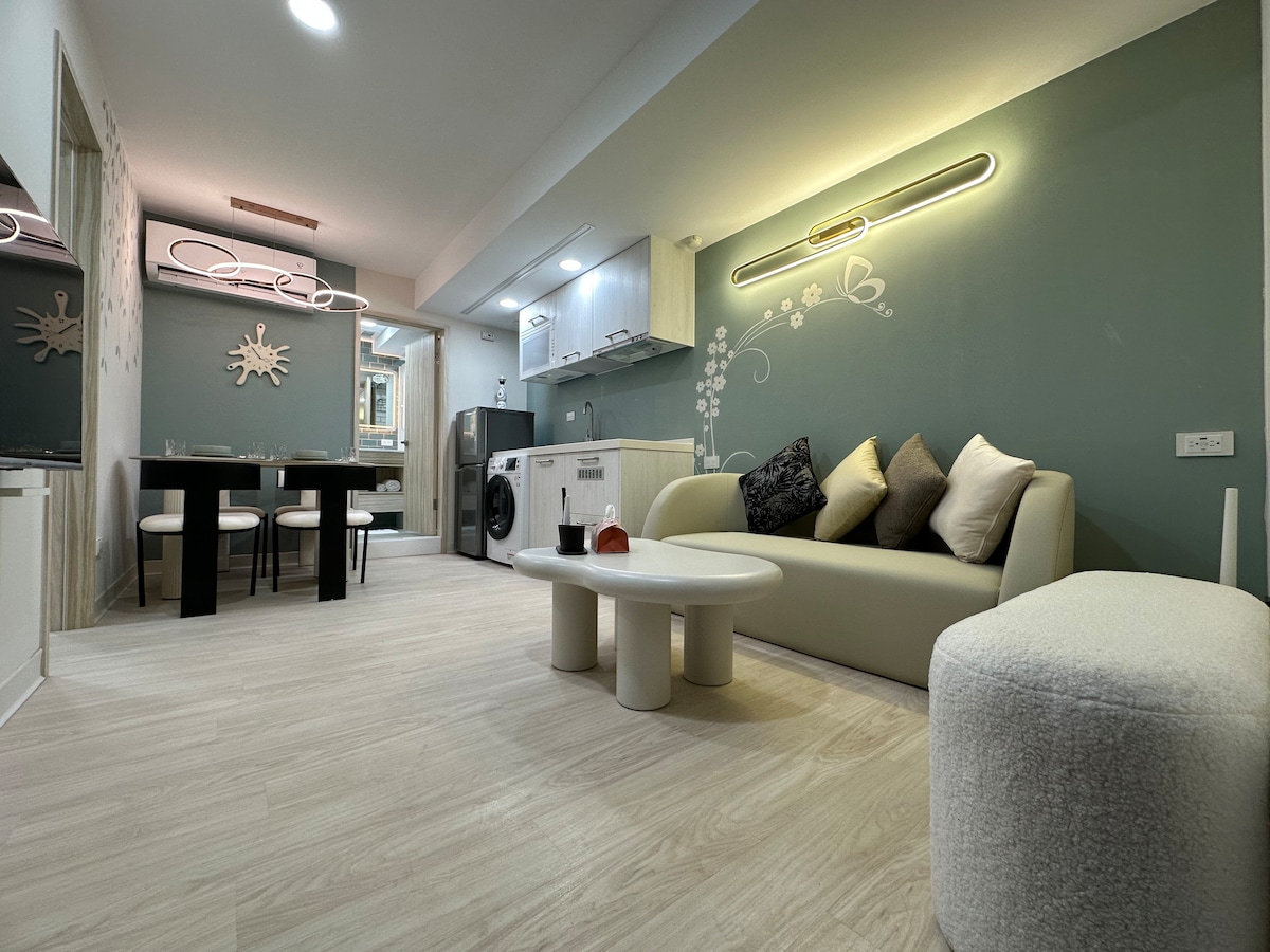 An open living area is shown with a light-colored sofa and a round coffee table. A dining table is set for four. Modern appliances in the kitchen are visible against a green accent wall with floral designs. Natural light fills the space through large windows.