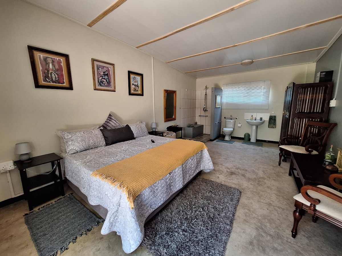 A spacious bedroom features a large bed adorned with a light yellow throw. Artwork hangs on the walls, while a rug lies underfoot. The en-suite bathroom is partially visible, showing modern fixtures and a shower. Soft lighting enhances the serene ambiance.