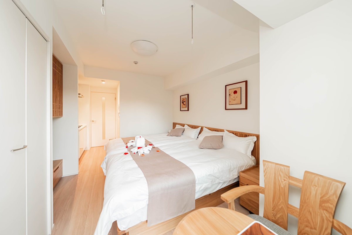 The room is furnished with a spacious double bed and a single bed, decorated with petals on the sheets. Light wood furniture includes a round table and two chairs. Two artworks featuring hearts adorn the white walls, contributing to a calming atmosphere.