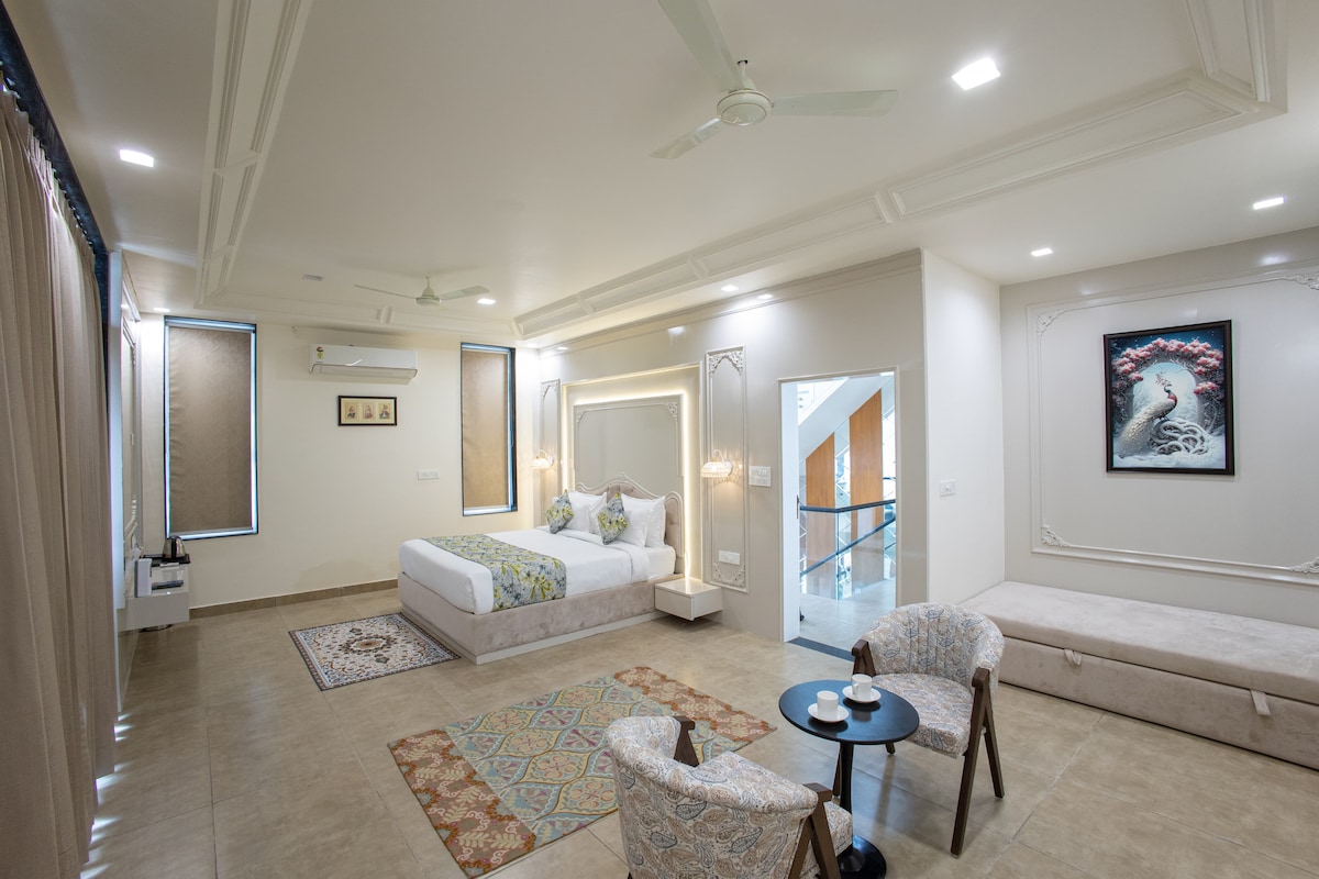 A spacious room is furnished with a large bed and a cozy, upholstered seating area. A small table is positioned between two stylish armchairs. Soft lighting frames the space, enhancing the elegant decor, while windows allow natural light to fill the room.
