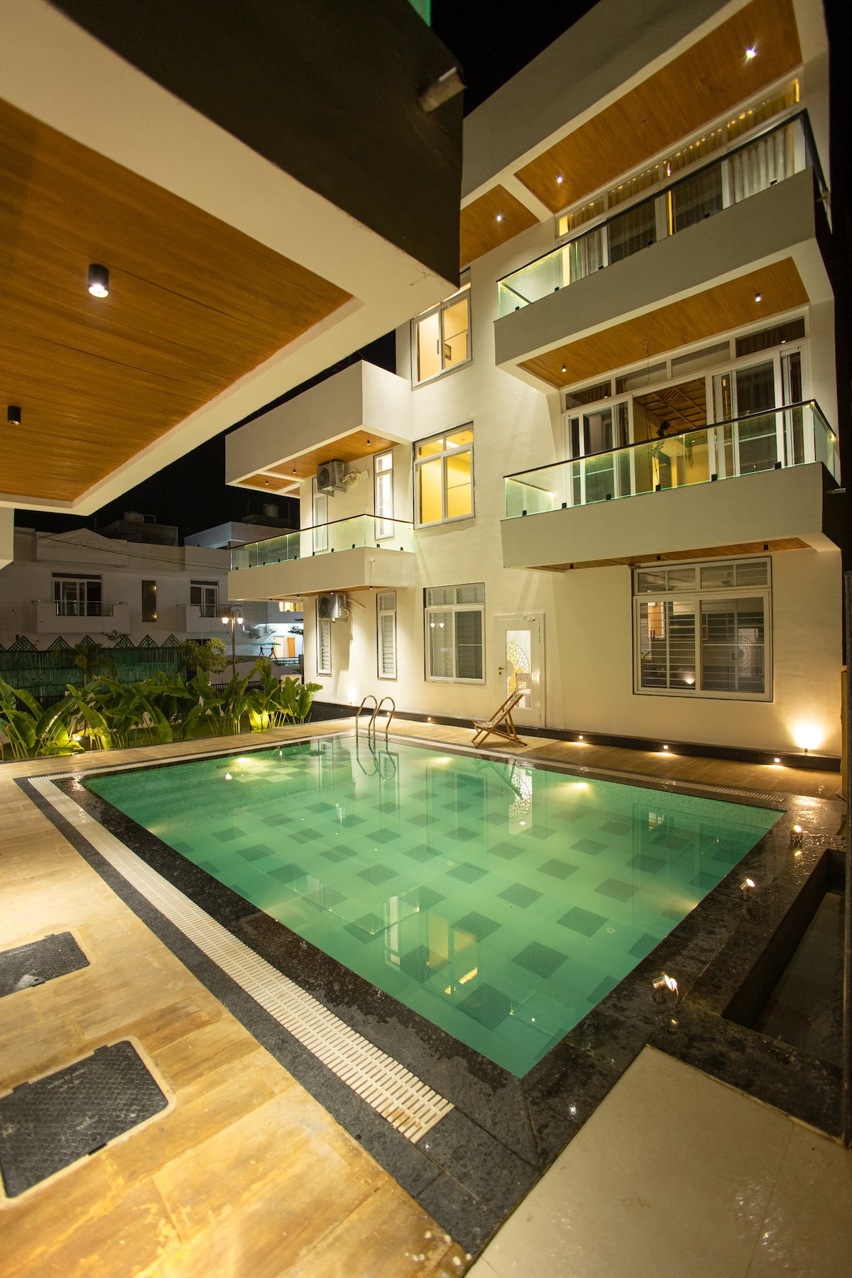 A modern building with multiple levels is depicted, featuring balconies and large glass windows. A clear swimming pool is visible in the foreground, surrounded by stylish stone tiles and illuminated at night, enhancing the structure's contemporary design.