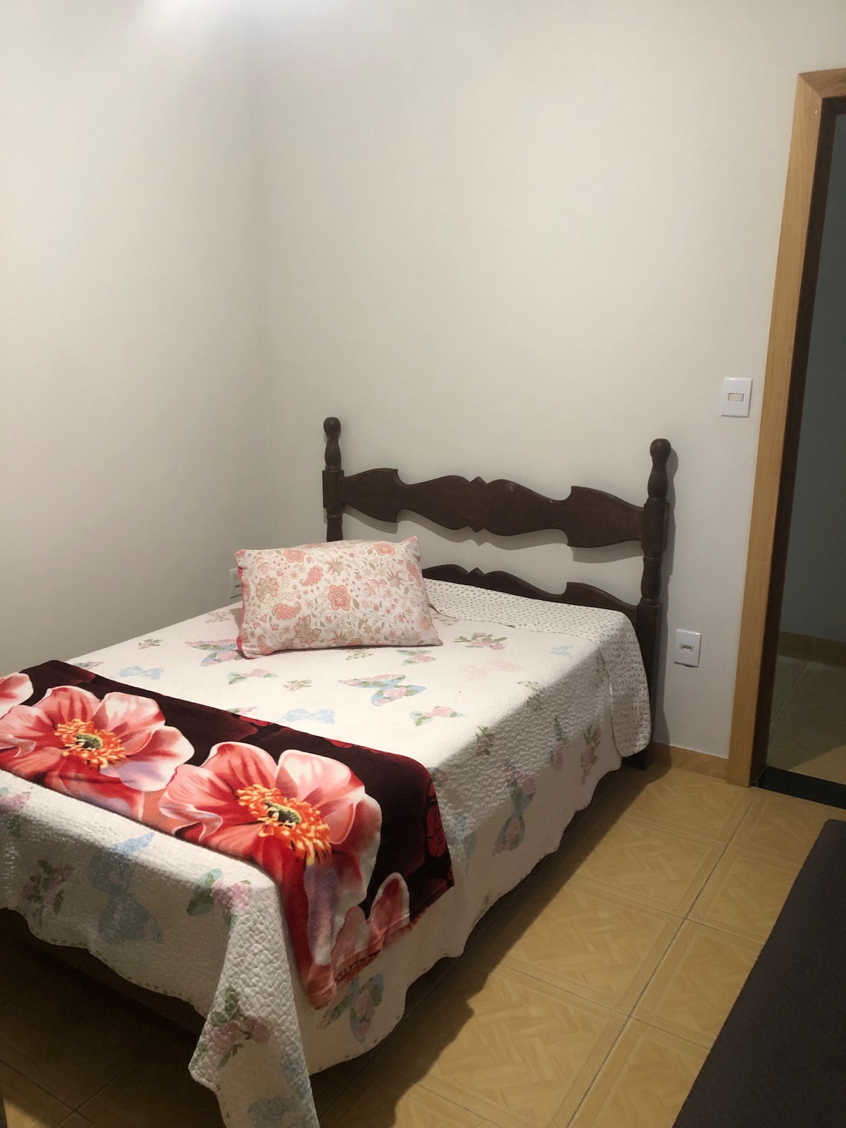 A cozy bedroom features a wooden bed with an intricate headboard. A floral pillow adds a touch of color. The bed is covered with a patterned quilt and a vibrant blanket, while light-colored walls and tiled flooring create a simple, clean backdrop.