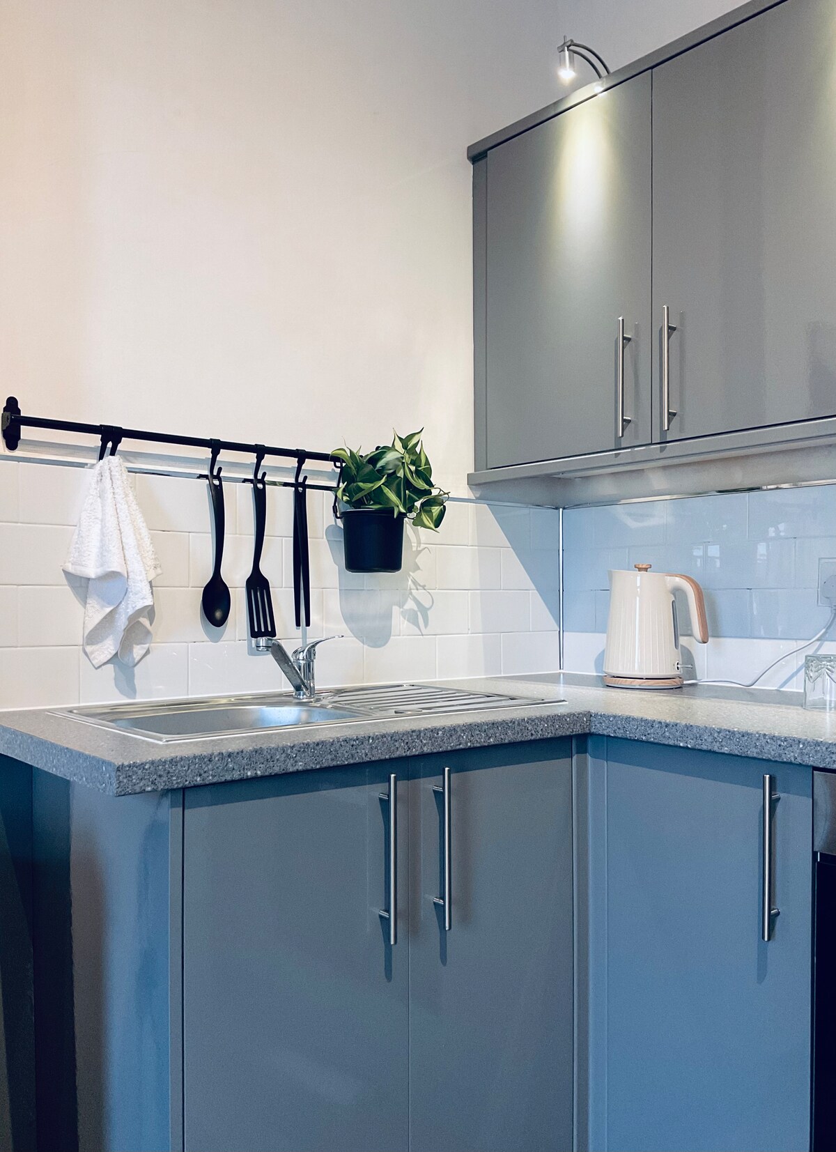 A modern kitchen is featured with sleek gray cabinetry and a smooth countertop. A wall-mounted rack holds cooking utensils, while a small potted plant adds a touch of greenery. A kettle is positioned beside a sink, with a bright light illuminating the workspace.