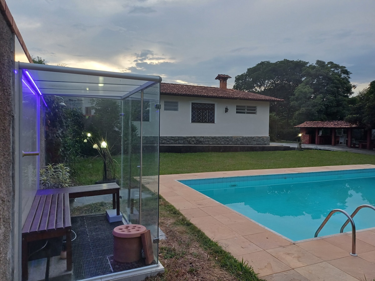 A clear view of an outdoor swimming pool is complemented by a well-maintained garden area. A gazebo made of glass features benches and lighting beside the pool, while a spacious house with a terracotta roof can be seen in the background.