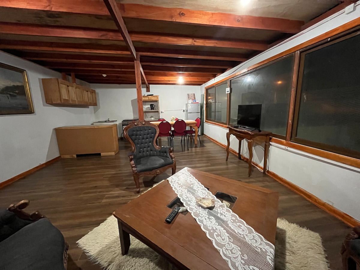 A spacious living area features warm wooden beams overhead, with a dining set of four red chairs visible. A comfortable seating arrangement, including two upholstered chairs, surrounds a central wooden coffee table. Large windows allow natural light to illuminate the space.