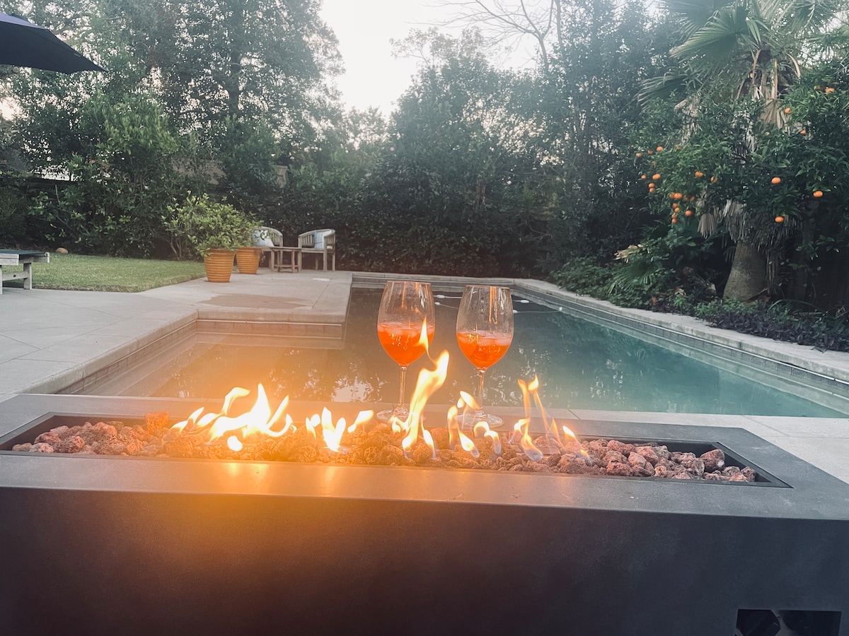 A fire table is displayed with flames dancing over decorative stones, positioned near a swimming pool. Two glasses filled with a vibrant drink are placed on the edge. Lush greenery surrounds the area, creating a serene outdoor space.