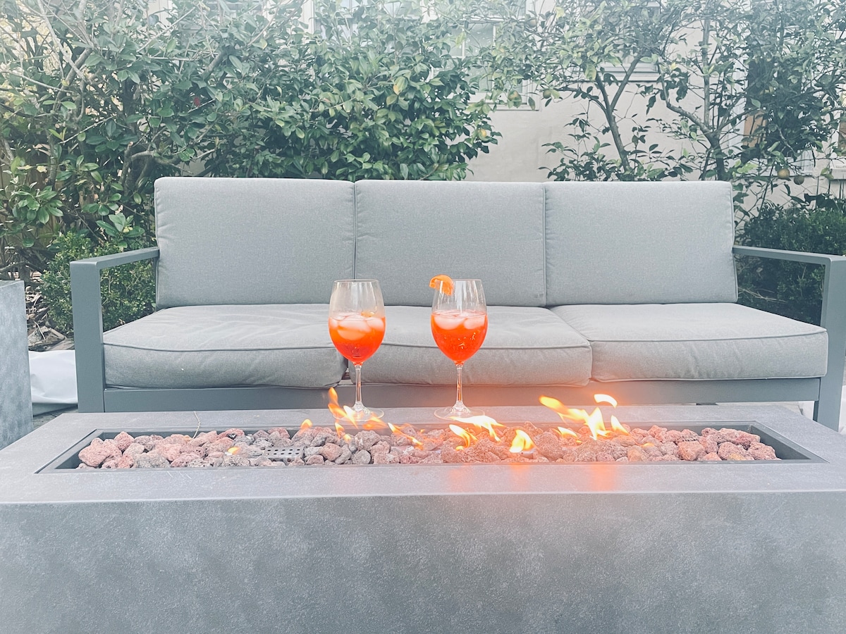 A cozy seating area features a gray outdoor sofa positioned near a fire table. Two glasses with vibrant orange drinks sit on the table, complemented by warming flames flickering amidst decorative stones.