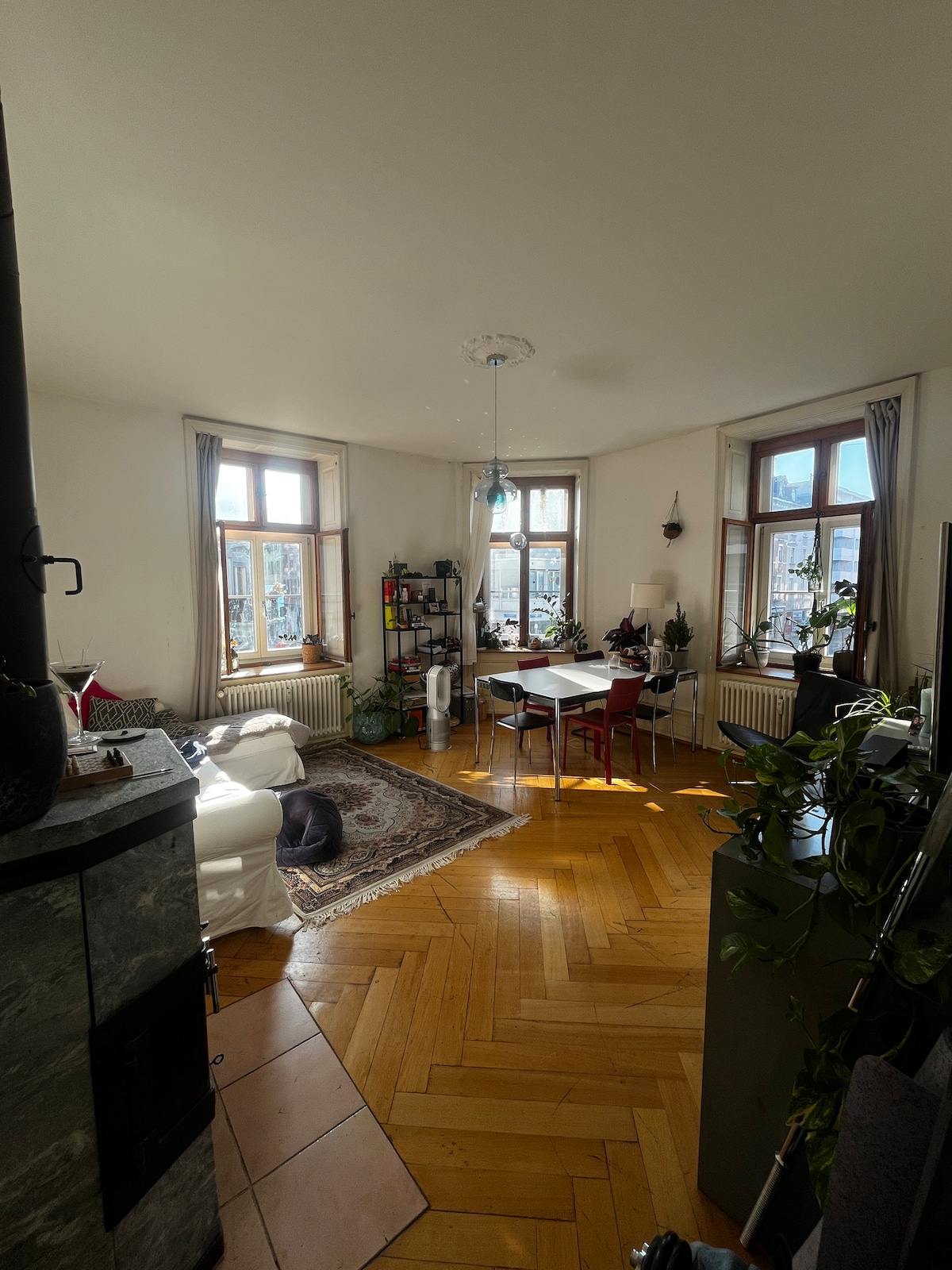 A spacious living area features large windows allowing natural light to illuminate the wooden parquet flooring. A dining table with chairs is positioned in the center, while a cozy seating area is arranged near a decorative stone fireplace. Decorative plants and a shelving unit add character to the space.