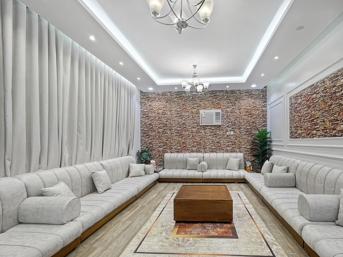 A spacious living area features a combination of soft, light-colored seating arranged in a U-shape. A wooden coffee table sits in the center, with decorative plants positioned in corners. The textured stone wall adds character, complemented by soft lighting from an elegant chandelier.