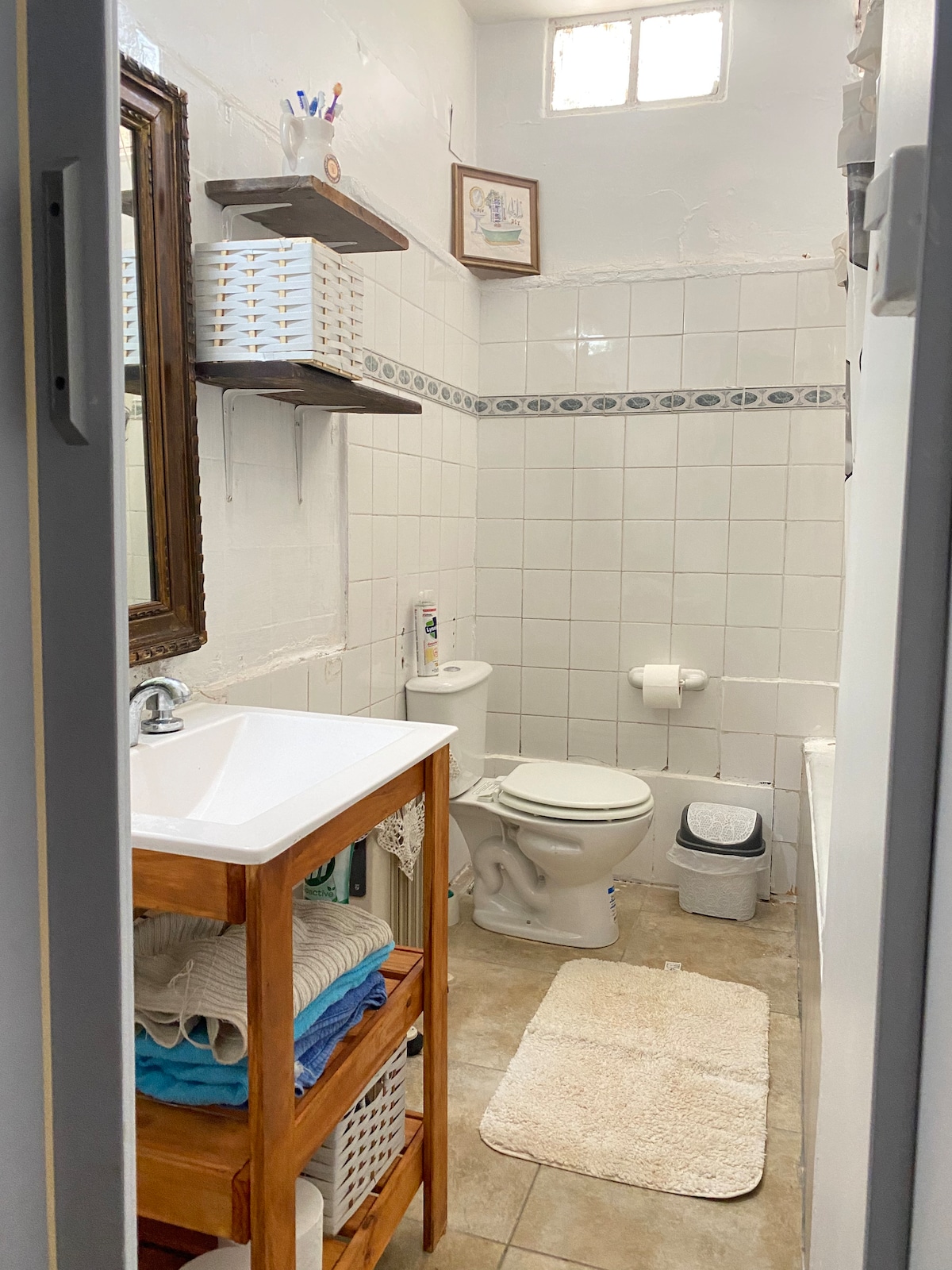 A compact bathroom features white tiled walls and a beige floor. A wooden vanity with storage holds neatly stacked towels, and a framed mirror reflects the space. Essential fixtures include a toilet and a small trash bin. Natural light enters through a small window.