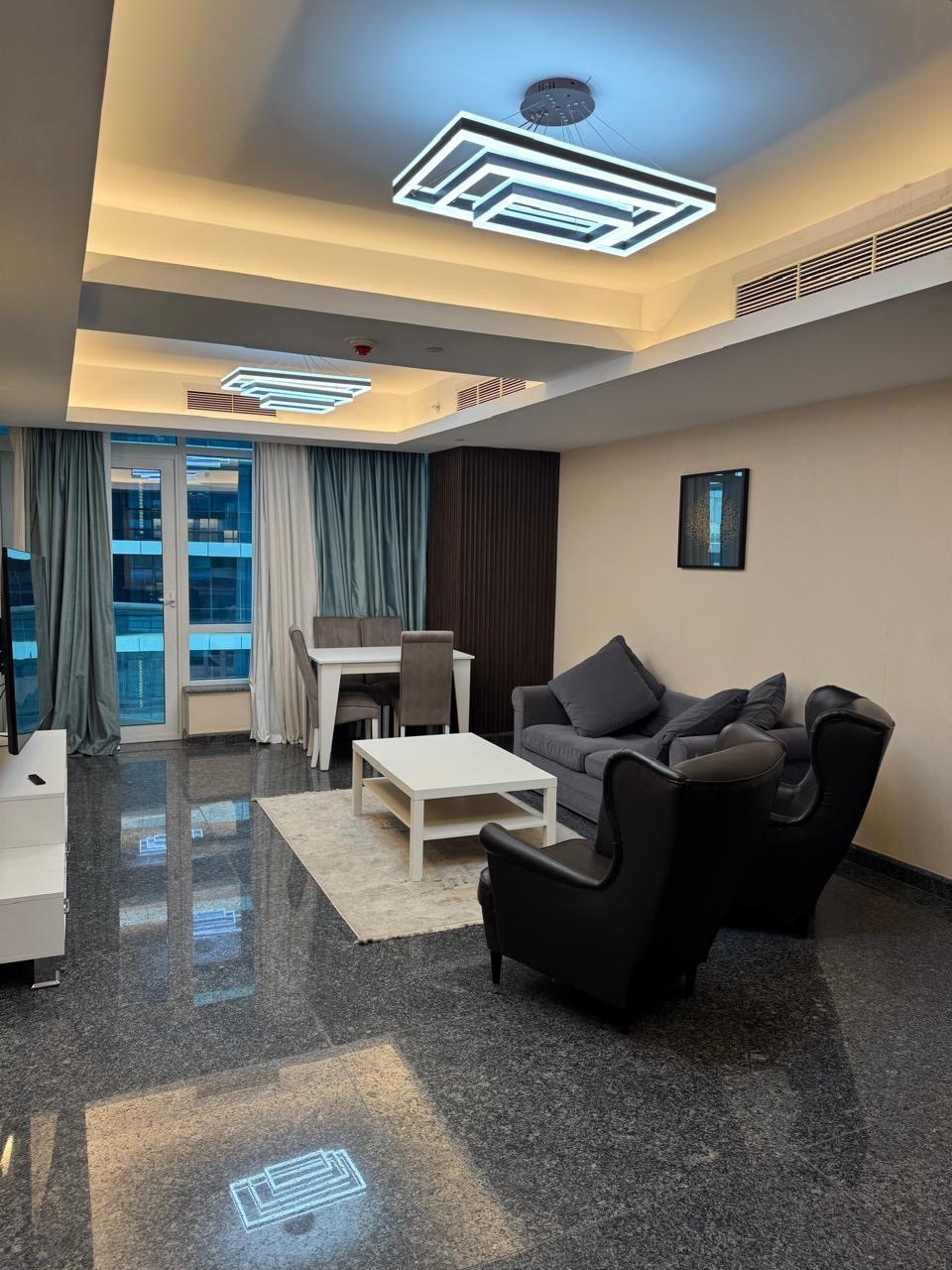 A modern living space features sleek, dark flooring and elegant lighting fixtures. A comfortable gray sofa is paired with armchairs, while a small dining table is visible near large floor-to-ceiling windows framed by light curtains. A minimalist wall frame hangs above the seating area.