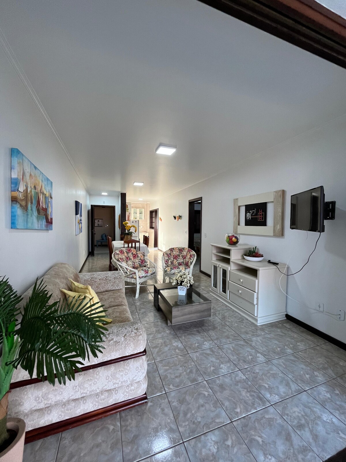 A well-lit living area is presented, featuring a light-colored sofa with decorative cushions and a glass coffee table. A television is mounted on the wall, and a cheerful painting adds color. A potted plant brings a touch of greenery, enhancing the inviting ambiance.
