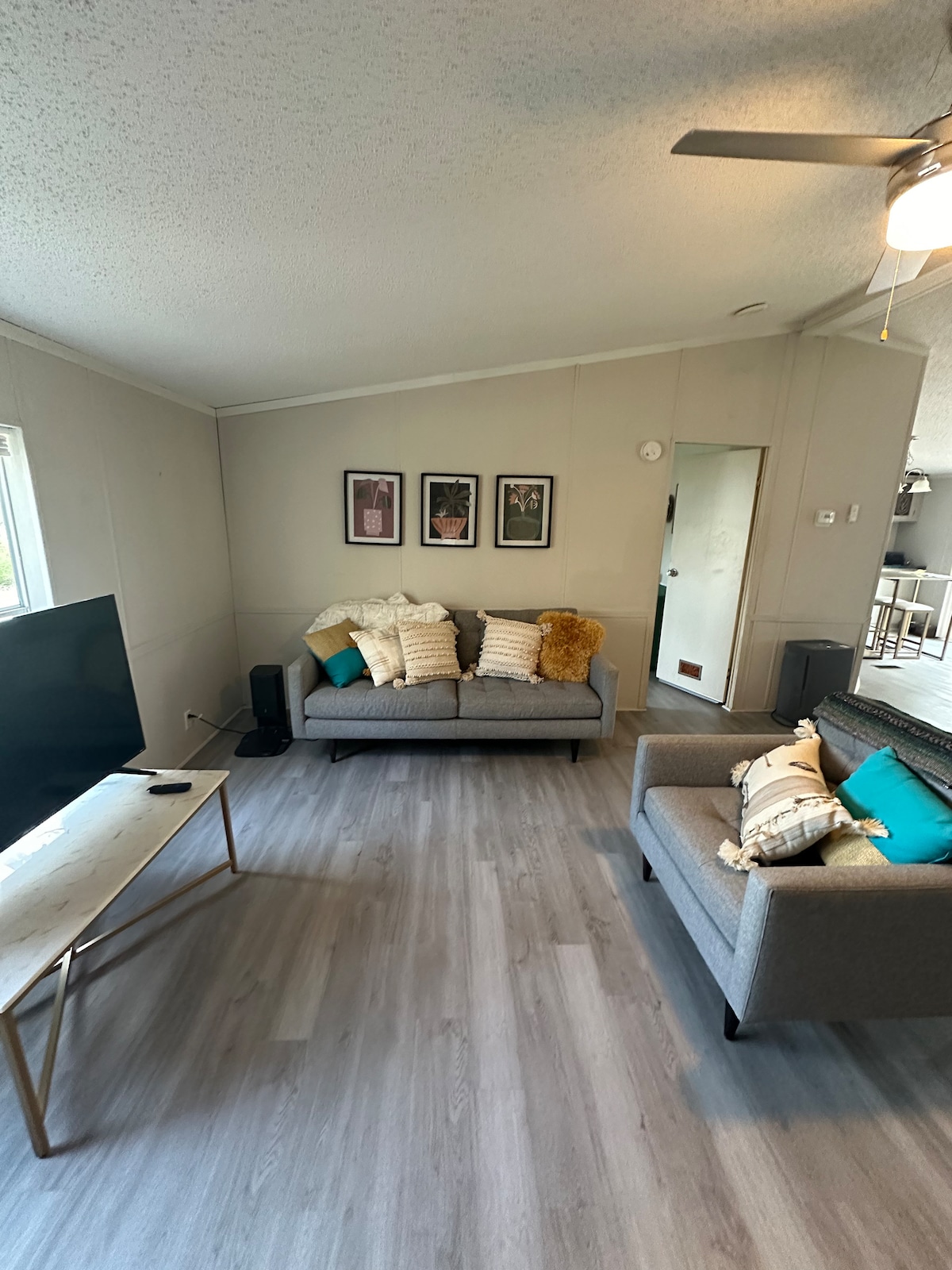 A cozy living area features two gray sofas, adorned with decorative pillows, positioned opposite a minimalist coffee table. A flat-screen television is displayed on a light wooden stand. The room is illuminated by natural light entering through large windows, highlighting the light-colored walls and floor.